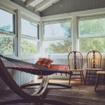 A bright sunroom with wooden floors features a red hammock with a pillow, two wooden chairs, and a tall laundry basket. Large windows on three sides let in ample sunlight and provide a view of green trees outside. The ceiling is wood-paneled.