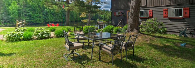 A backyard scene with a glass-top patio table and six cushioned chairs on a grassy lawn. The area is shaded by a large tree and surrounded by greenery. Red Adirondack chairs are placed near a lake in the background beside a rustic wooden house with red shutters.