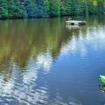 A serene lake surrounded by lush green trees reflects the sky and clouds. A green kayak rests on a blue dock in the foreground, while a yellow and a red canoe float on the lake. A wooden platform is seen further in the lake.