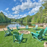 A picturesque lakeside scene with a circle of green Adirondack chairs surrounding a stone fire pit on a lush grassy area. The lake is calm, surrounded by dense forest under a bright blue sky with scattered clouds. Canoes rest on the shore in the background.