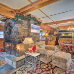 A cozy rustic living room decorated for the holidays with a large stone fireplace adorned with garlands. There's a patterned armchair, matching ottoman, and a small table with a lamp. In the background, there's a dining area with a wooden table and chairs.