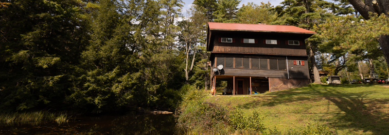 A two-story wooden cabin with a red roof sits beside a calm pond, surrounded by tall trees. The cabin features a screened porch on the lower level and multiple windows. The grass lawn in front is lit by the afternoon sun, and a satellite dish is attached to the cabin.