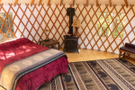 A cozy yurt interior features a bed with a red quilt, a small wood stove, a bench, and a patterned rug. The yurt has wooden lattice walls, two windows allowing natural light, and a wooden door. The floor is wooden, and the space is warmly lit and inviting.