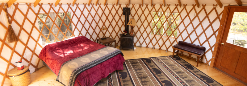 A cozy yurt interior features a bed with a red quilt, a small wood stove, a bench, and a patterned rug. The yurt has wooden lattice walls, two windows allowing natural light, and a wooden door. The floor is wooden, and the space is warmly lit and inviting.