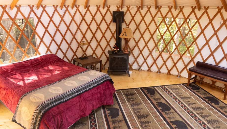 A cozy yurt interior features a bed with a red quilt, a small wood stove, a bench, and a patterned rug. The yurt has wooden lattice walls, two windows allowing natural light, and a wooden door. The floor is wooden, and the space is warmly lit and inviting.