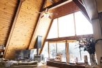 A cozy A-frame cabin interior with exposed wooden beams and high ceilings. The living area features a large window allowing natural light to fill the space, a comfortable sofa, a TV, and rustic decor, including a vase with flowers on a wooden counter.