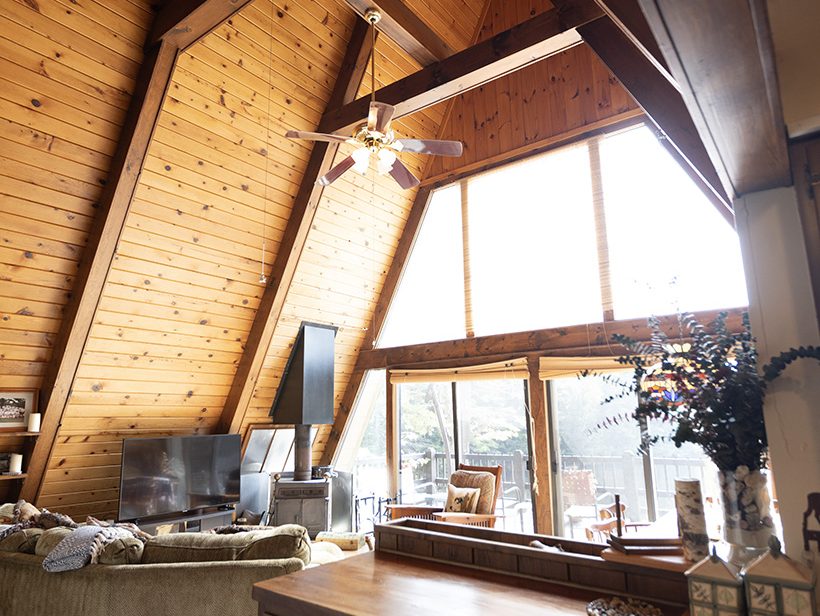 A cozy A-frame cabin interior with exposed wooden beams and high ceilings. The living area features a large window allowing natural light to fill the space, a comfortable sofa, a TV, and rustic decor, including a vase with flowers on a wooden counter.