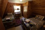 A cozy room with wooden walls featuring two single beds adorned with plaid and animal-patterned bedding. A colorful rug lies between the beds. A window with light curtains lets in natural light next to a small dresser with a lamp and a mirror above it.