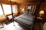 A cozy wooden bedroom features a large bed with a patterned black, white, and red quilt. Two bedside tables with lamps are on either side of the bed. A window with white curtains allows natural light to fill the room. A small dresser and chair are placed near the window.