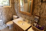 A bathroom featuring a wooden vanity with a sink and a large mirror, illuminated by multiple lights. The walls are covered with tree-themed wallpaper. A toilet is next to the vanity. Various toiletries are organized in a basket on a small shelf nearby.