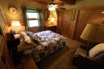 A cozy bedroom with wooden paneling and a ceiling fan. The bed is covered with a colorful patchwork quilt featuring nautical designs. Warm lighting comes from table lamps on bedside tables. There is a rocking chair with a floor lamp beside it in the corner.