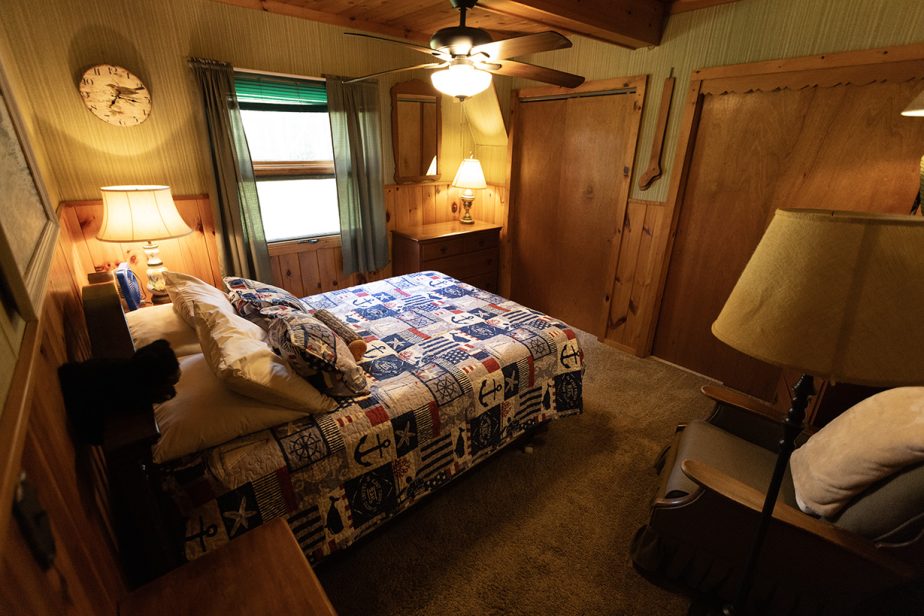 A cozy bedroom with wooden paneling and a ceiling fan. The bed is covered with a colorful patchwork quilt featuring nautical designs. Warm lighting comes from table lamps on bedside tables. There is a rocking chair with a floor lamp beside it in the corner.