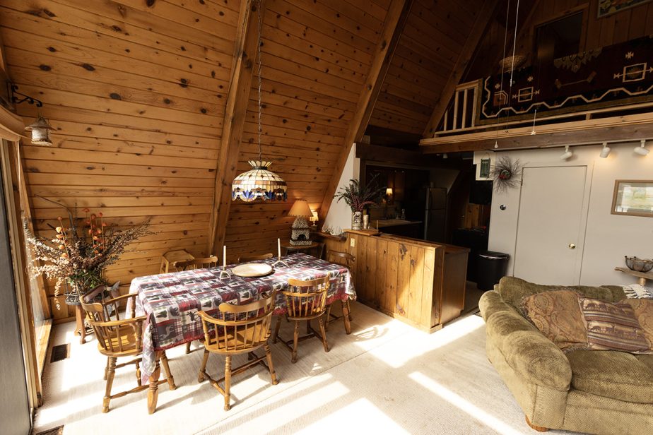 A cozy A-frame cabin interior with wood-paneled walls and high ceilings. The dining area features a table with a patterned tablecloth and wooden chairs. A comfortable sitting area is on the right, and a loft with rustic decor is visible above the kitchen.