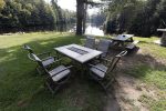 A serene outdoor setting featuring a square table with four chairs on a grassy area by a calm lake. Nearby, there is a wooden picnic table. The background includes trees and the lake reflecting the tranquil environment.