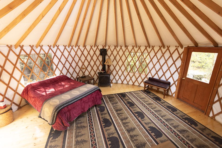 Interior of a cozy yurt with wooden lattice walls and a round roof. The room features a bed with a burgundy comforter, patterned rugs with bear designs, a small wood stove, and a wooden door with a window. Two small square windows provide natural light.