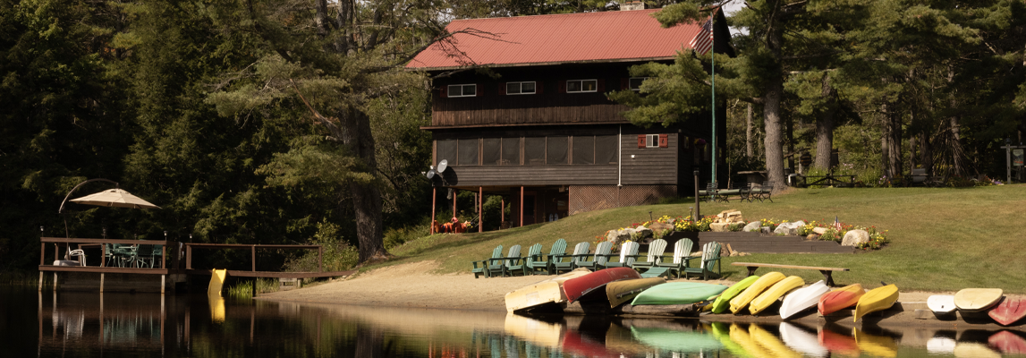 A serene lakeside scene featuring a rustic wooden cabin with a red roof. Outside, several colorful kayaks and canoes are lined upside down on the shore. The area has chairs set up for relaxation, surrounded by lush green trees and a calm body of water.