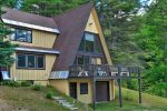 A large, rustic A-frame house surrounded by tall pine trees. The house has yellow siding with brown trim and a steep roof. A wooden deck extends from the second floor, supported by tall posts, and there are grassy patches around the property.