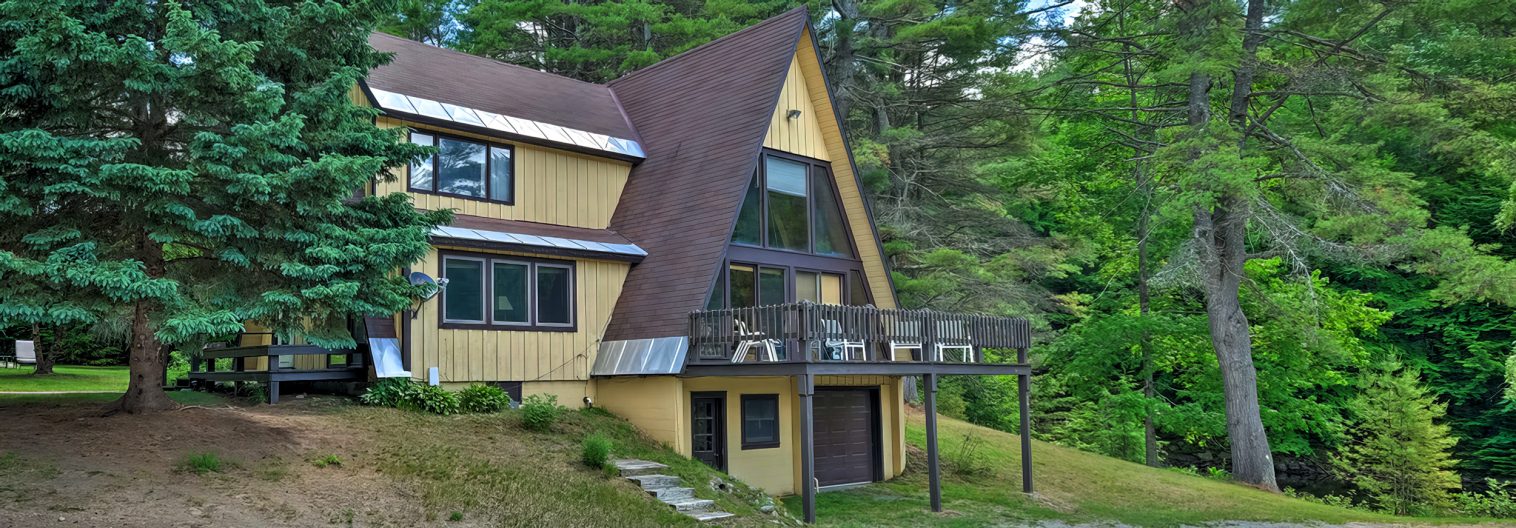 A large, rustic A-frame house surrounded by tall pine trees. The house has yellow siding with brown trim and a steep roof. A wooden deck extends from the second floor, supported by tall posts, and there are grassy patches around the property.