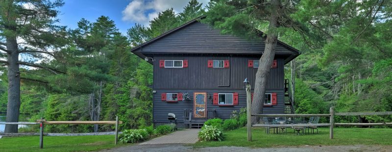 A two-story black house with red window shutters, surrounded by lush green trees and bushes. There's an outdoor seating area with a table and chairs on a wooden deck beside the house. The sky is partly cloudy. A body of water is visible on the left side.