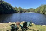 Four green Adirondack chairs surround a fire pit, facing a tranquil lake with clear blue water. The lake is bordered by dense, green trees under a bright blue sky. There are a couple of houses visible on the distant shoreline.