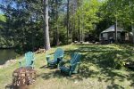 A scenic outdoor setting features four green Adirondack chairs arranged around a fire pit on a grassy lawn. In the background, a round tent-like structure stands near a dense forest of tall trees on a bright sunny day. A calm body of water is visible to the left.