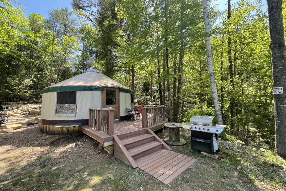 A yurt with a green roof sits in a wooded area. The yurt has a wooden deck with stairs, a small table with chairs, and a BBQ grill on the ground nearby. Lush green trees surround the area, and sunlight filters through the foliage.