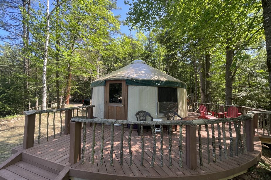 A yurt with a beige canvas exterior and wooden door is surrounded by a spacious, elevated wooden deck. The deck has rustic railings made from natural branches and is furnished with plastic chairs, set amidst a green, leafy forest under a clear blue sky.