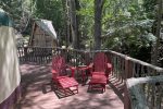 A wooden deck in a forested area features two red Adirondack chairs with footrests and a small table between them. The deck railing surrounds the space, and a small rustic wooden structure is seen in the background among the trees.