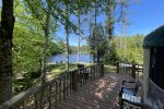 A wooden deck with railing overlooks a serene lake surrounded by lush trees. The deck has chairs and tables, and the sun shines brightly, casting a peaceful atmosphere. In the background, dense forest lines the lake's edge under a clear blue sky.