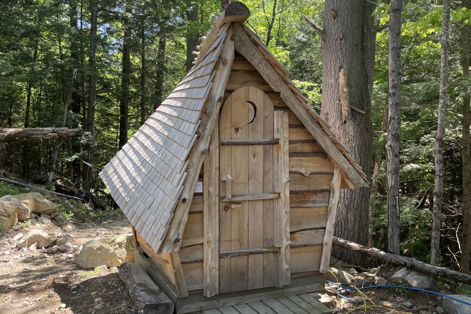 A small, rustic wooden cabin with a triangular roof stands in a wooded area. The cabin has a weathered door with a crescent moon cutout, and is situated at the edge of a wooden deck amidst tall trees and scattered rocks. Sunlight filters through the leaves above.