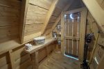 A small wooden cabin interior with roughly finished wooden walls and ceiling. A narrow door with gaps is at the end. A shelf to the left holds various items, including a roll of toilet paper. There is a vacuum cleaner and a broom leaning against the wall.