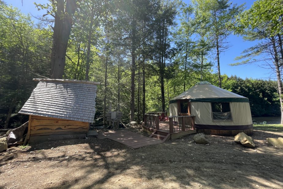 A round yurt with a wooden deck sits in a sunny, forested area. To the left, there is a small wooden structure with a slanted roof. Trees and a blue sky can be seen in the background, casting dappled shadows on the ground.