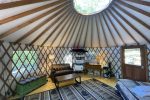 Interior view of a cozy yurt with wooden lattice walls and a circular skylight. The yurt is furnished with a couch, coffee table, green chest, bookshelf, and a small kitchenette area. A patterned rug covers the wooden floor and soft light filters in from a window and door.