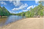 A tranquil lake surrounded by dense green trees under a partly cloudy sky. Small boats are moored on the calm water, and a sandy beach extends from the shoreline. A wooden deck with seating overlooks the lake, providing a scenic spot for relaxation.