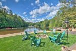 A scenic lakeside area with a circle of green Adirondack chairs surrounding a fire pit on a grassy lawn. The calm lake is bordered by tall trees under a bright blue sky with white clouds. Canoes and a small dock are visible along the shoreline.