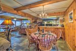 A cozy wood-paneled dining area features a round table with a checkered tablecloth and wooden chairs. Above hangs an antler chandelier. The background includes a kitchen bar with stools, rustic decor, and various decorations like signs and garlands.