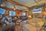 A cozy living room with brown leather couches and an armchair arranged around a coffee table. The room features wood paneling, a fireplace with stone surround, a wall-mounted TV, and windows letting in natural light. The space is decorated with a festive touch.