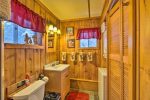 A cozy, rustic bathroom with wooden walls and red curtains. It features a white toilet, a small wooden vanity with a white sink, and a washer and dryer hidden behind a louvered door. Several small framed pictures adorn the walls and a red mat adds color to the floor.