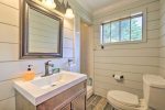 A bright bathroom with light wood panel walls featuring a large mirror above a white sink with a wooden base. It includes a modern faucet, soap dispenser, towel rack, toilet, and a window above the toilet. A shower stall is partially visible in the background.