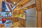 A cozy kitchen with wooden cabinets, a stainless steel refrigerator, an electric stove with a red kettle, and a sink under a window. A dishwasher is next to the sink. The kitchen has an overhead wooden beam and a decorative plate collection on the counter.