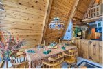 A cozy dining area in a wooden cabin features a table set for a meal with six wooden chairs. The room has cedar plank walls and an A-frame ceiling. A colorful stained glass chandelier hangs above the table adorned with a patterned tablecloth, candles, and foliage decor.