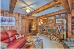 A cozy room with wooden walls and a ceiling fan. A red leather couch faces a TV. There's Native American decor, horse paintings, and a variety of vintage signs on the walls. A dog lies on the carpet, and a staircase is visible in the background.