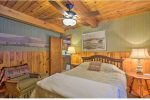 A cozy bedroom with wood-paneled walls and ceiling. The room features a bed with a white and beige comforter, a wooden headboard, a bedside table with a lamp, and an armchair with a patterned blanket. A ceiling fan and framed pictures adorn the room.