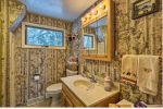 A small rustic bathroom features woodland-themed decor, including wallpaper with tall tree designs. A wooden vanity with a sink and mirror is adorned with lit bulbs. A window provides natural light. Towels with forest animal patterns hang beside the sink.