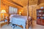 A cozy bedroom with wooden paneled walls and ceiling featuring a four-poster bed with gray and white bedding. There are nightstands with lamps on each side of the bed, a step stool at the foot of the bed, and a wooden bookcase with various items to the right.