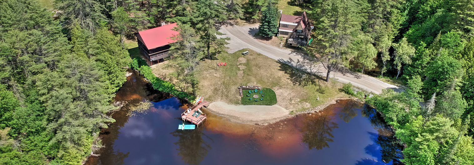 Aerial view of a lakeside area with two buildings surrounded by dense trees. A boat is docked by the lake, and there's a small pier and lawn space with chairs and a firepit. A dirt road runs between the buildings and the lawn area.