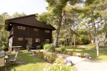 A rustic wooden cabin surrounded by trees, featuring a well-maintained garden with colorful flowers and a small pathway leading to the front entrance. A painted sign stands near the pathway, and a picnic area with benches is visible to the left.