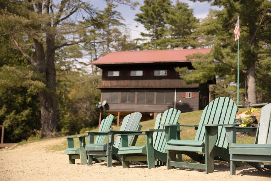 A rustic wooden cabin with a red roof is surrounded by trees. In the foreground, several green Adirondack chairs are arranged on a sandy area, facing the cabin. An American flag is visible on a flagpole near the cabin. The scene is summery and tranquil.