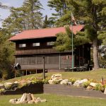 A rustic wooden cabin with a red metal roof stands on a grassy lawn surrounded by trees. An American flag is raised on a flagpole by the cabin. In the foreground, there is a landscaped area with flowers, large rocks, and a circular fire pit.