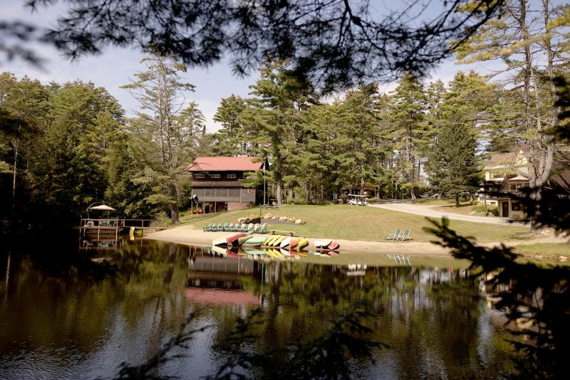 A serene lakeside scene features a rustic lodge surrounded by tall pine trees. Several colorful kayaks and paddleboats are lined up on the shore. The calm water reflects the peaceful environment under a clear sky, with Adirondack chairs set in the distance.